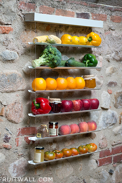 Frutero de pared colgante sobre pared de piedra, contiene 2 mazorcas de maiz, 3 limones, 1 pimiento amarillo, 1 brocoli, 2 calabacines 1 pimiento verde, 5 naranjas, 1 pimiento rojo, 4 manzanas, 4 melocotones, 6 tomates 4 botes de mermelada y un bore de pimienta en grano.