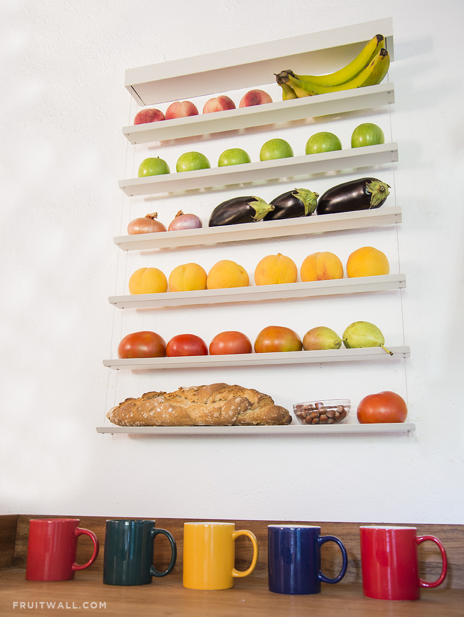 Fruitwall, los mejores fruteros modernos y de diseño para la cocina, colocado sobre pared blanca. esta visto desde abajo, esta lleno de frutas, verduras, una barra de pan y un cuenco con avellanas, debajo hay 5 tazas de diferentes colores.